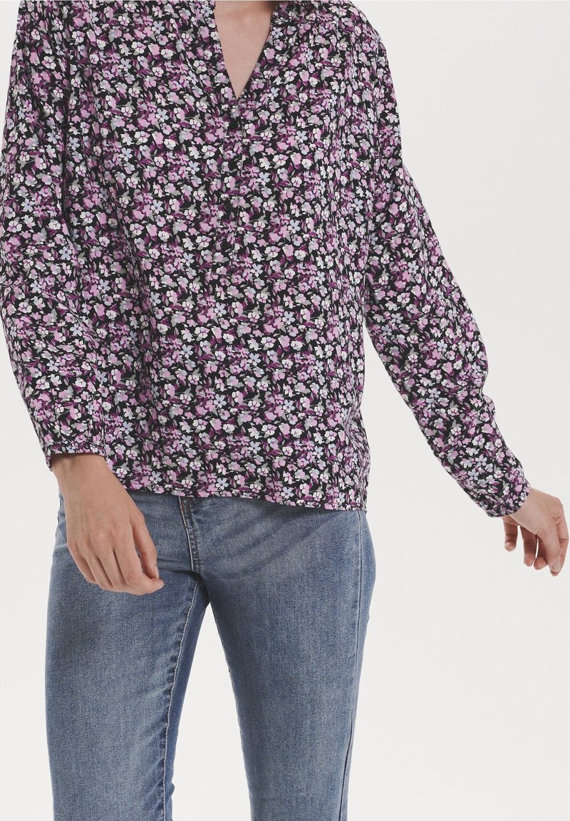 Blouse fleurie en tissu noir avec des fleurs roses et blanches, dotée de manches longues bouffantes et d'un décolleté en V. Associée à un jean bleu.