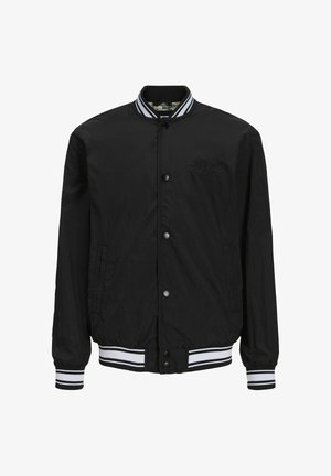 Veste bomber noire avec des poignets rayés blancs, pull beige, shorts en denim et baskets avec des accents noirs et des détails blancs.