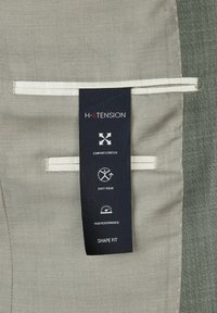 H-XTENSION kledinglabel op een gestructureerde grijze stof. Bevat iconen voor "Comfort Stretch," "Easy Wear," en "High Performance." Vorm pasvorm aangegeven.