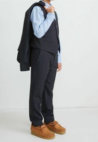 Navy three-piece suit with a fitted vest, light blue shirt, and brown suede moccasin shoes. Jacket draped over the arm.