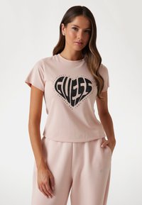 Vaaleanpunainen puuvillapaita lyhyillä hihoilla, jossa on musta sydämenmuotoinen "GUESS"-logo, jota koristavat sirotteet, yhdistettynä yhteensopiviin vaaleisiin housuihin.