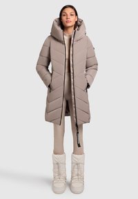 Polstret, lang beige frakke med chevron-syning, aftagelig hætte, sidelommer og lynlås-lukning, parret med hvide isolerede støvler.