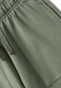 Zbliżenie na oliwkowe bawełniane spodnie z elastycznym paskiem w talii, boczną kieszenią i detalem sznurka.