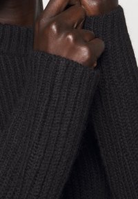 Schwarzer Strickpullover mit gerippten Bündchen und einer weichen, strukturierten Oberfläche. Nahaufnahme von Händen, die den Stoff greifen.