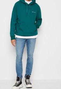 Sudadera verde azulado con bolsillo canguro y cordones, llevada sobre una camiseta blanca. Combinada con jeans de un azul claro y zapatillas altas negras.