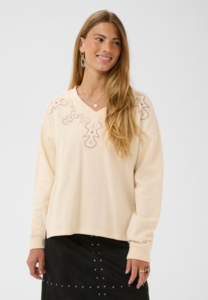 Jeune femme aux longs cheveux blonds portant un pull crème avec des motifs brodés et une jupe noire à clous métalliques, souriante et regardant sur le côté.