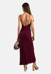 Burgunderfarbenes Spaghettiträgerkleid mit tiefem Rückenausschnitt, figurbetonter Silhouette und fließendem Saum. Kombiniert mit einer metallisch goldenen Handtasche und klobigen Absätzen.
