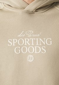 Béžová mikina s jemnou texturou, zdobená vystouplým bílým logem s nápisem "Les Deux SPORTING GOODS" v elegantním fontu. Nastavitelné šňůrky na kapuci.