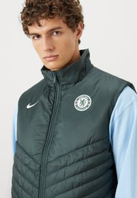Grøn polstret vest med høj krave, lynlåsbeslåning og Chelsea Football Club-logo. Har et struktureret, quiltet mønster og Nike-logo.