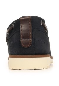 Vista posteriore di una scarpa in camoscio nero con linguetta in pelle marrone e suola in gomma beige su sfondo bianco.