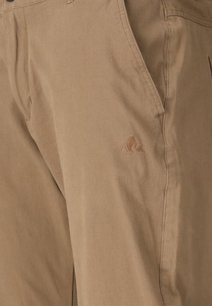 Pantalon beige avec une poche latérale et un logo triangulaire subtil cousu près de la poche, mettant en valeur la texture du tissu décontracté et les détails de couture.