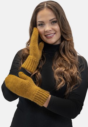 Manoplas de color mostaza, tejidas, con puños acanalados, que muestran una textura suave y forma redondeada, sostenidas por una persona con una camiseta negra.