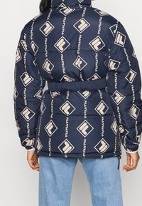 Granatowa puchowa kurtka z beżowym wzorem w logo na całej powierzchni. Posiada pasek w talii, wysoki kołnierz oraz elastyczne mankiety zapewniające pewne dopasowanie.