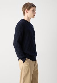 Maglione lavorato a maglia blu navy con motivo a rombi, caratterizzato da un collo rotondo e polsini a coste, abbinato a un pantalone beige.