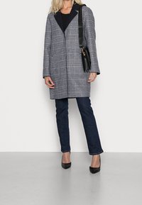 Una donna che indossa un cappotto a quadri bianco e nero, jeans scuri, tacchi neri e porta una borsa a tracolla nera su uno sfondo semplice.