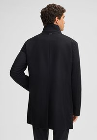 Cappotto nero in lana con colletto alto, design strutturato e taglio dritto. Presenta uno stile minimalista e una texture liscia sul retro.