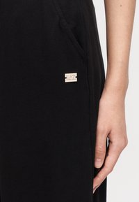 Δεξί χέρι που ακουμπά δίπλα σε ένα μαύρο ένδυμα με μικρό χρυσό λογότυπο EA7 Emporio Armani κοντά στην τσέπη.