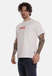 T-shirt bianca in cotone con maniche corte, caratterizzata da un logo grafico rosso sul petto. Abbinata a jeans scuri, che mostrano una vestibilità rilassata.