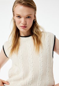 Strickweste ohne Ärmel in Creme mit schwarzen Akzenten. Verfügt über ein Kabelstrickmuster und einen runden Halsausschnitt. Weiche Textur.