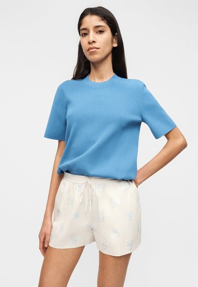 Jeune femme aux longs cheveux bruns portant un haut bleu à manches courtes et un short blanc à cordon avec motif floral, debout la main sur la hanche.