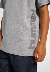 Grå bomulls-T-shirt med en stor grå tryckt "hummel"-logotyp vertikalt längs vänster sida. Enkel design, korta ärmar, avslappnad passform.