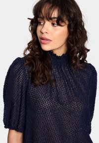 Tengerészkék, áttetsző blúz textúrázott, hímzett felülettel, magas nyakkal és puffos ujjal, lágy, folyékony vonalvezetéssel.