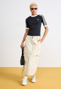 Svart kortermet topp med hvite detaljer og tre striper, parret med en kremfarget cargo-skjørt og hvite sneakers med grønne detaljer.