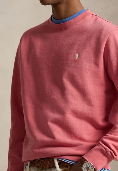 Άνδρας που φοράει κοραλλί ροζ φούτερ με μικρό κεντητό λογότυπο, μπλε πουκάμισο από κάτω, καφετί πλεγμένη ζώνη και ρολόι χειρός με χαρακτήρα της Disney.