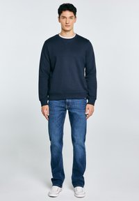 Jonge man staand, gekleed in een donkerblauwe trui, blauwe spijkerbroek en witte schoenen, tegen een effen lichte achtergrond.