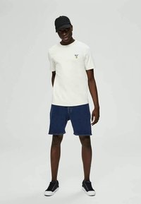 Man wearing black cap, white short-sleeve t-shirt with small graphic, blue denim shorts, and black sneakers standing against a plain background.