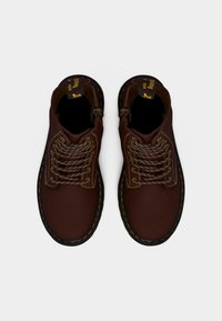 Ζευγάρι καφέ δερμάτινων μποτών με κορδόνια και κίτρινη ραφή, με μαύρες ταινίες έλξης με λογότυπο, φωτογραφημένα από πάνω σε λευκό φόντο.