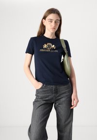 T-shirt bleu marine avec un design de logo doré, associé à un jean large gris et un sac à épaule vert avec une sangle.