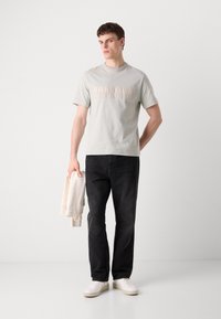 Světle šedé tričko s krátkým rukávem a nápisem "BARBOUR INTERNATIONAL" béžovým písmem, v kombinaci s černými džínami a bílými teniskami, držící béžovou mikinu.