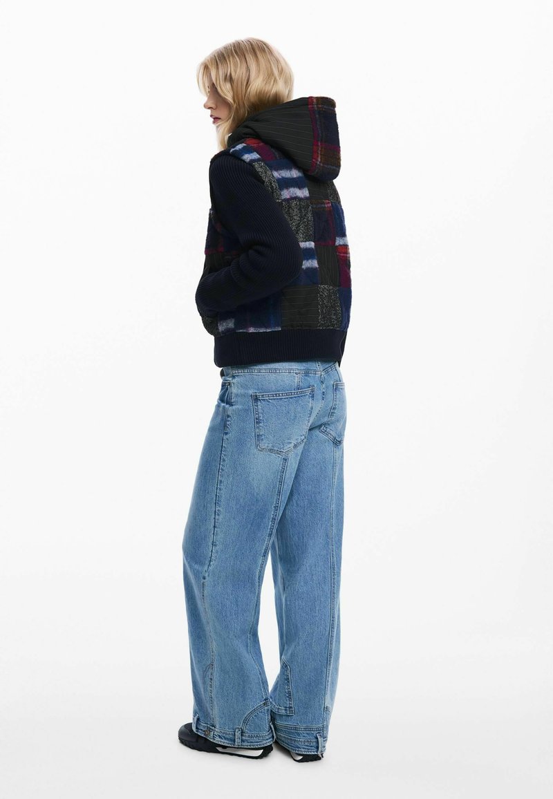 Giacca a hood patchwork nera e multicolore con una texture a costine, abbinata a jeans a gamba larga blu chiaro e scarpe nere.