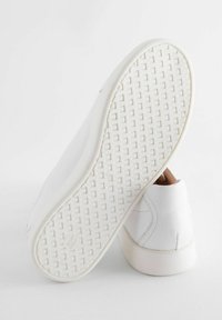 Baskets blanches en cuir avec une finition texturée, semelle en caoutchouc plate dotée d'un motif en grille, et détails de surpiqûre subtils sur la tige.