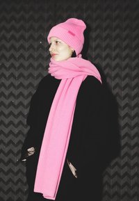 Căciulă roz tricotată și eșarfă asortată, ambele cu textură nervurată. Stilizate cu o haină neagră pe un fundal negru texturat.