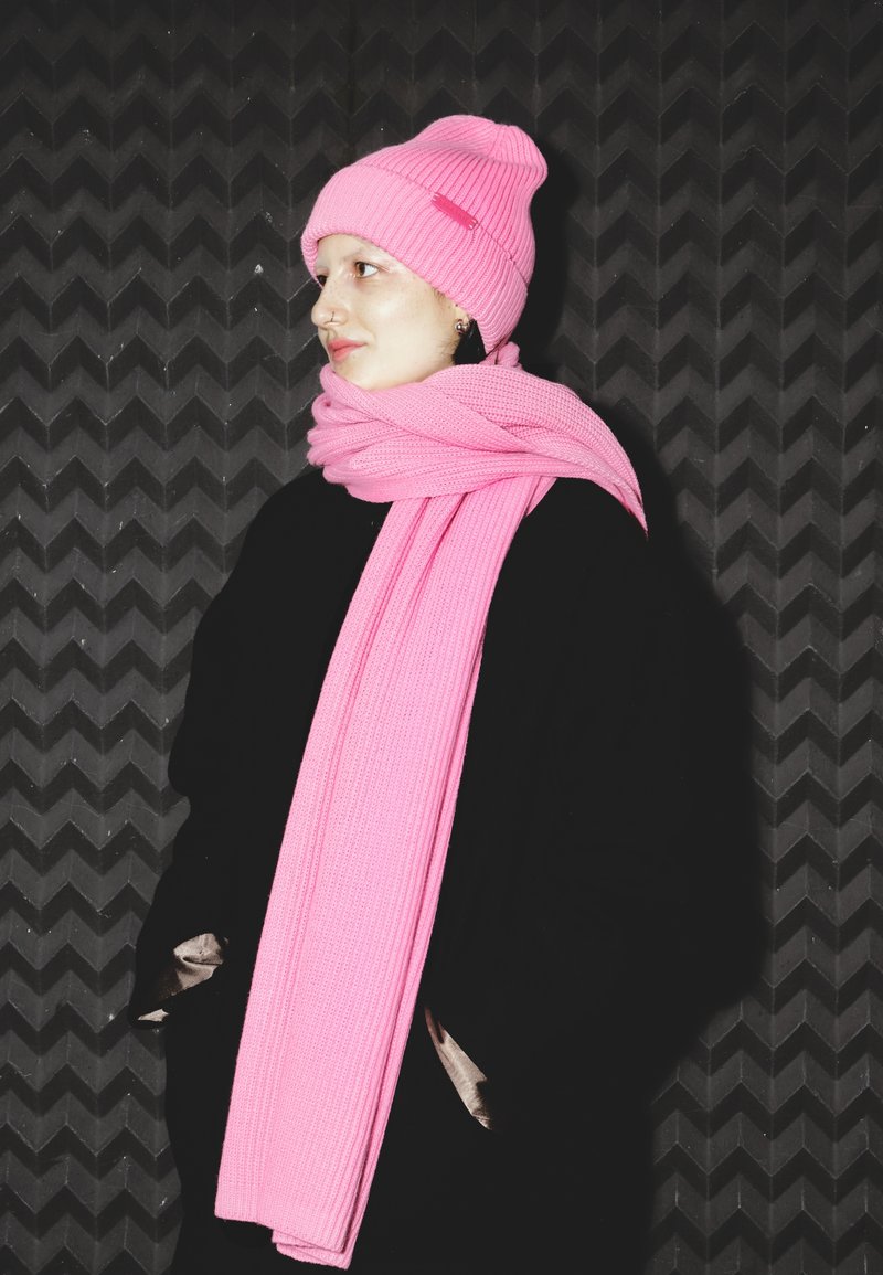 Căciulă roz tricotată și eșarfă asortată, ambele cu textură nervurată. Stilizate cu o haină neagră pe un fundal negru texturat.