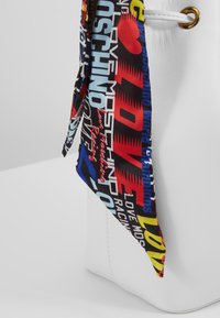 Vitt läderhandväska med en färgglad mönstrad scarf med textaccenter i rött, blått, gult och svart, fäst högst upp.