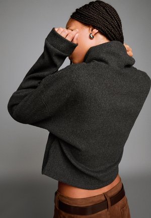 Temno siva rebrasta turtleneck pulover z ohlapnim krojem, dolgimi rokavi in visokim ovratnikom, v kombinaciji z rjavimi suknjičnimi hlačami.