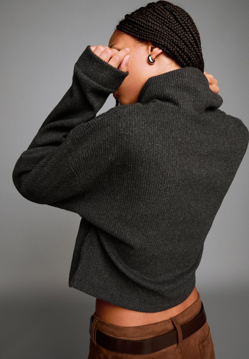 Temno siva rebrasta turtleneck pulover z ohlapnim krojem, dolgimi rokavi in visokim ovratnikom, v kombinaciji z rjavimi suknjičnimi hlačami.
