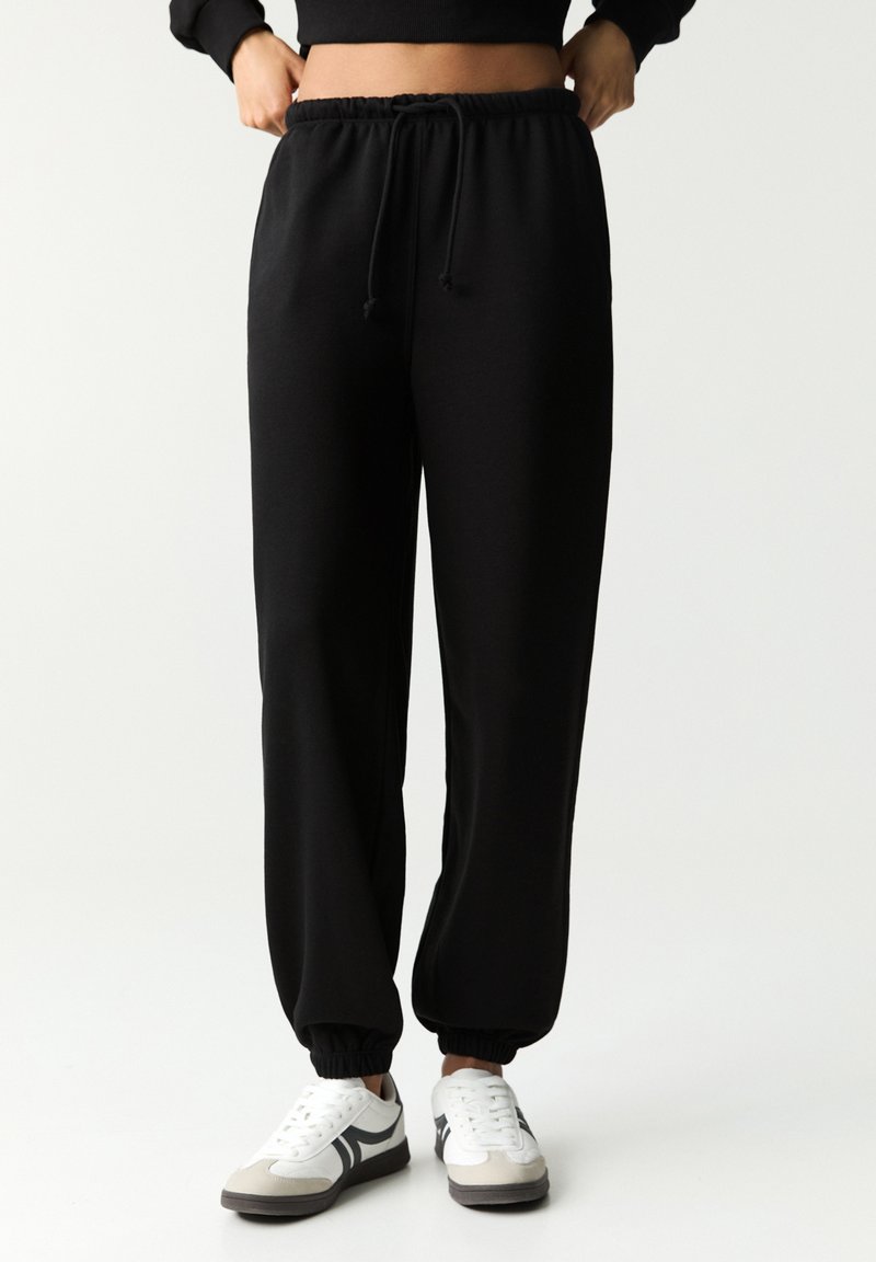 Pantaloni de jogging negri cu talie elastică, șnur ajustabil și manșete înguste. Asortați cu adidași albi și gri.
