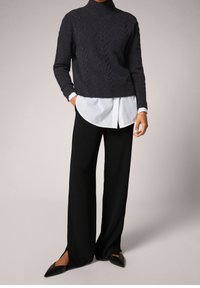 Personne portant un pull col roulé en maille torsadée gris foncé, chemise blanche en dessous, pantalon large noir avec fentes, et chaussures plates pointues noires.