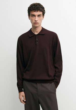 Junger Mann trägt ein dunkelbraunes langärmeliges Poloshirt und braune Hose und steht vor einem schlichten weißen Hintergrund.