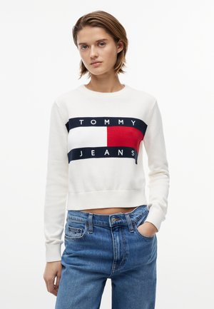 Ung person med kort hår iført en hvid Tommy Jeans-sweater og blå jeans, stående med den ene hånd i lommen mod en hvid baggrund.