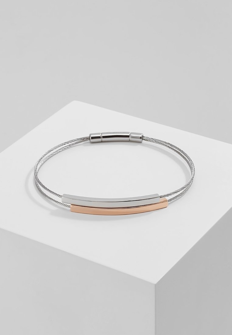 Bracelet fin en fil d'argent avec des barres courbées en argent et or rose reposant sur un piédestal blanc sur fond gris.