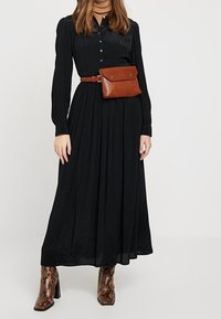 Abito nero a maniche lunghe con bottoni, gonna plissettata e borsa a cintura in pelle marrone. Abbinato a stivali con tacco in stampa pitone. Tessuto dalla texture liscia.