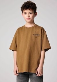Brūns kokvilnas T-krekls ar īsām piedurknēm, kuram kreisajā pusē ir melns drukāts logotips. Valkāts ar pelēkiem džinsiem. Vienkāršs dizains, brīvs piegriezums.