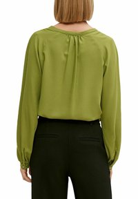 Blusa verde a maniche lunghe con scollo rotondo, tessuto strutturato, pieghe posteriori e polsini con bottoni, abbinata a pantaloni neri sartoriali.