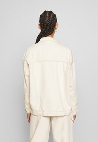 Krämfärgad denimjacka med avslappnad passform, synlig sömnad och knäppte ärmslut. Modellen bär matchande krämfärgade byxor.