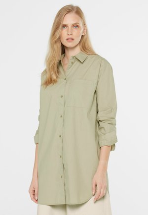 Blonde Frau trägt langes, hellolivgrünes Hemd mit aufgekrempelten Ärmeln, steht vor einem einfachen weißen Hintergrund.